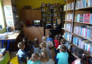 Dzieci siedzą pomiędzy regałami książek, oglądają film edukacyjny.